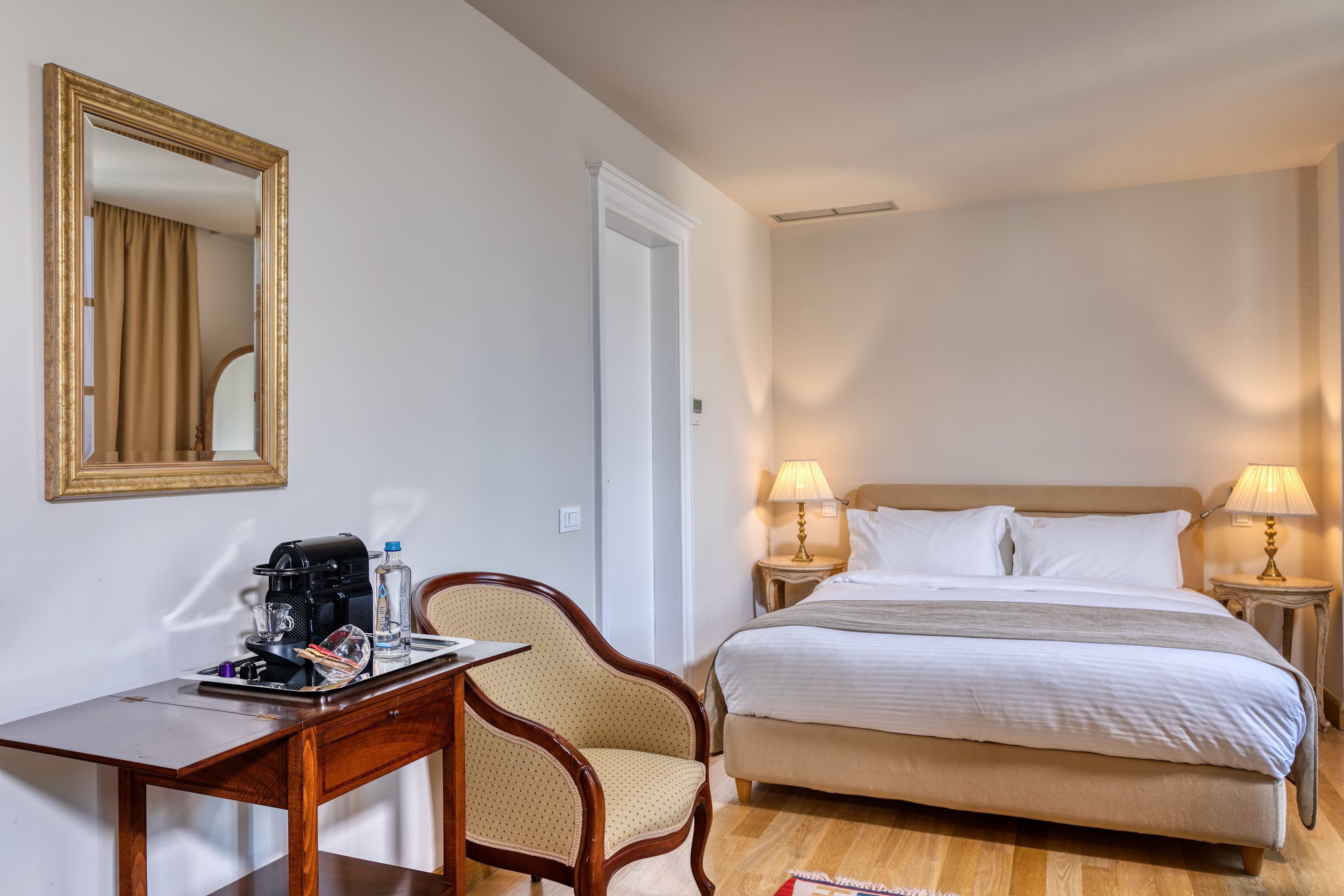 A hotel room with a bed, two bedside lamps, a wooden desk with a coffee maker, a chair, and a mirror on the wall.