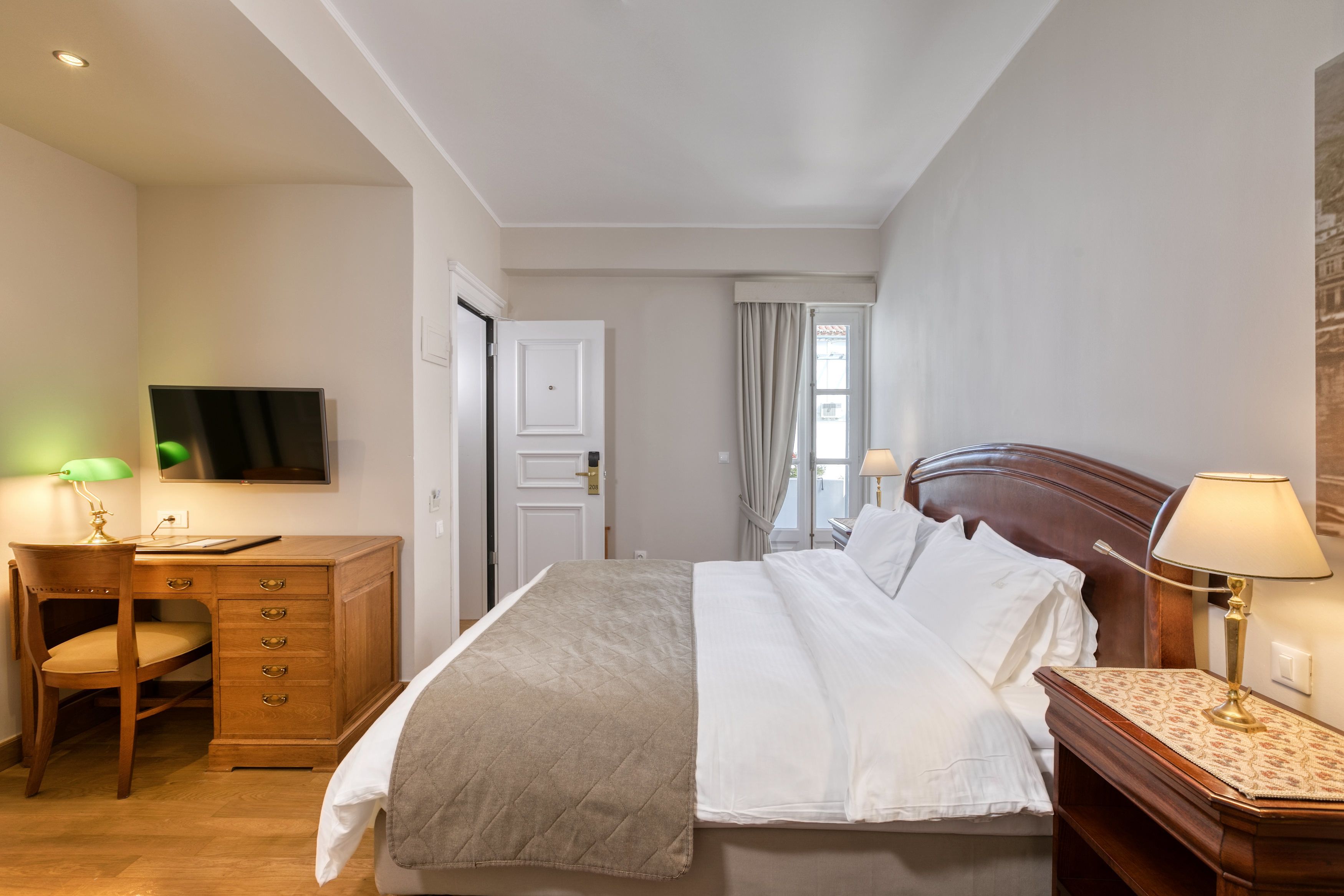 Quarto de hotel com cama, escrivaninha com cadeira, televisão e mesa de cabeceira com abajur.