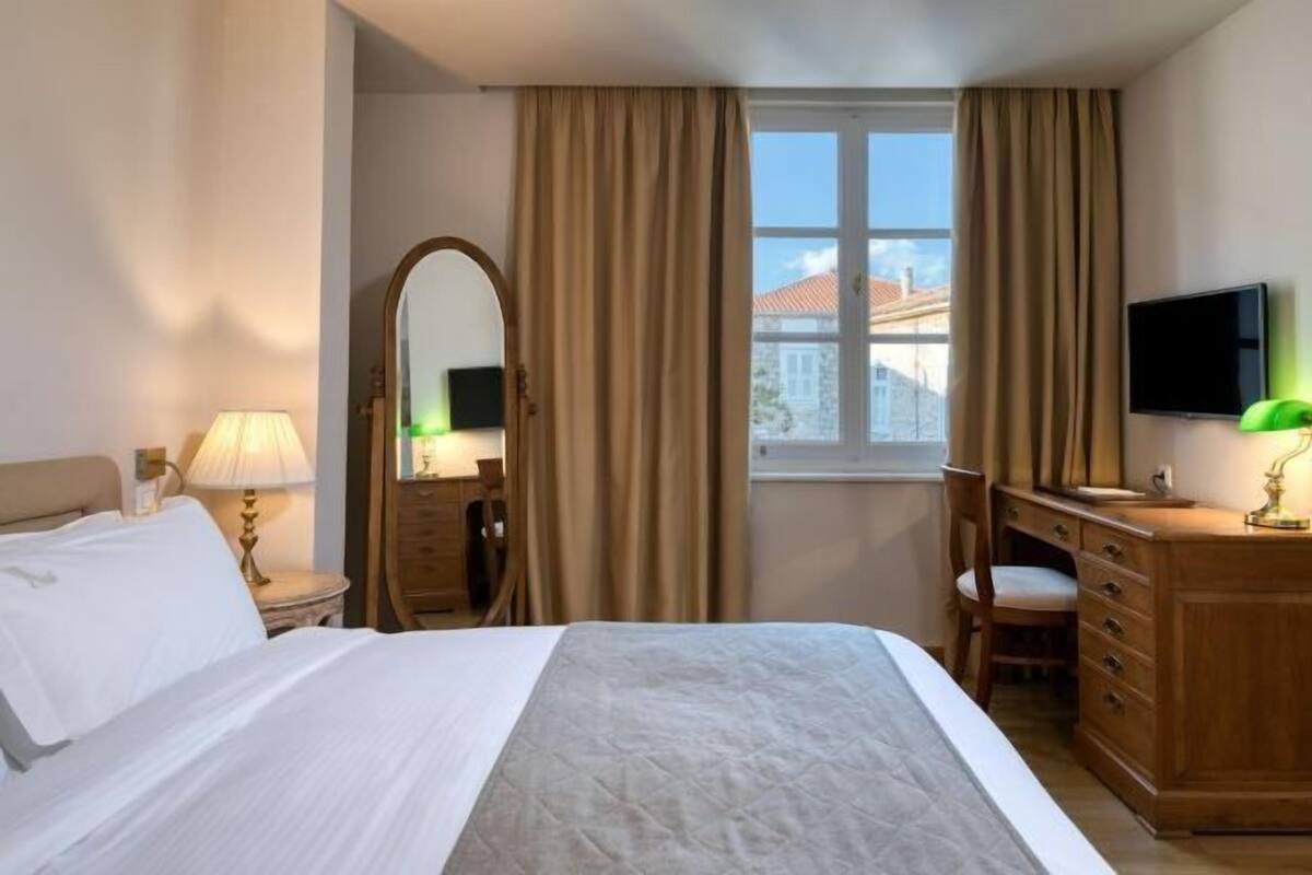 Habitación de hotel con cama, escritorio, silla, televisión, espejo y ventana con cortinas.