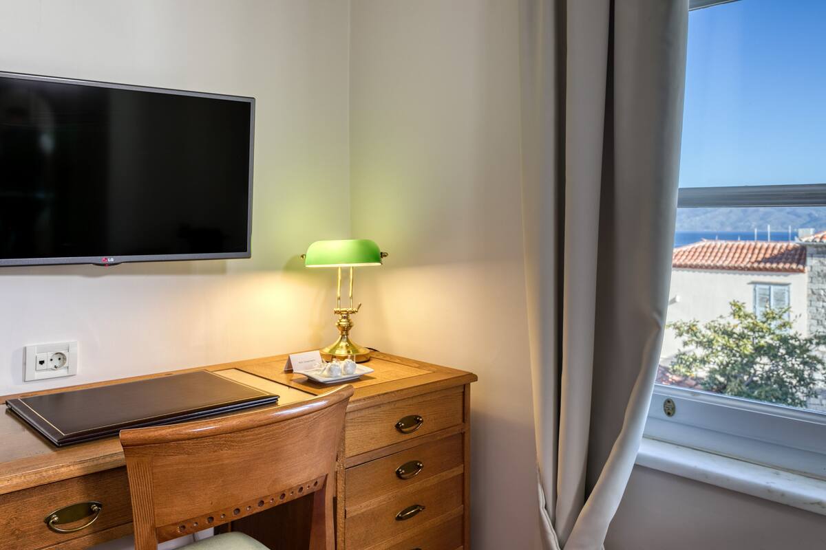Una habitación de hotel con un escritorio de madera, un televisor de pantalla plana, una lámpara de escritorio verde y una ventana con vistas a edificios y árboles.