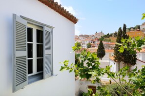 A white building with a window featuring horizontal blinds, a view of a town with red-tiled roofs, and green foliage in the foreground.