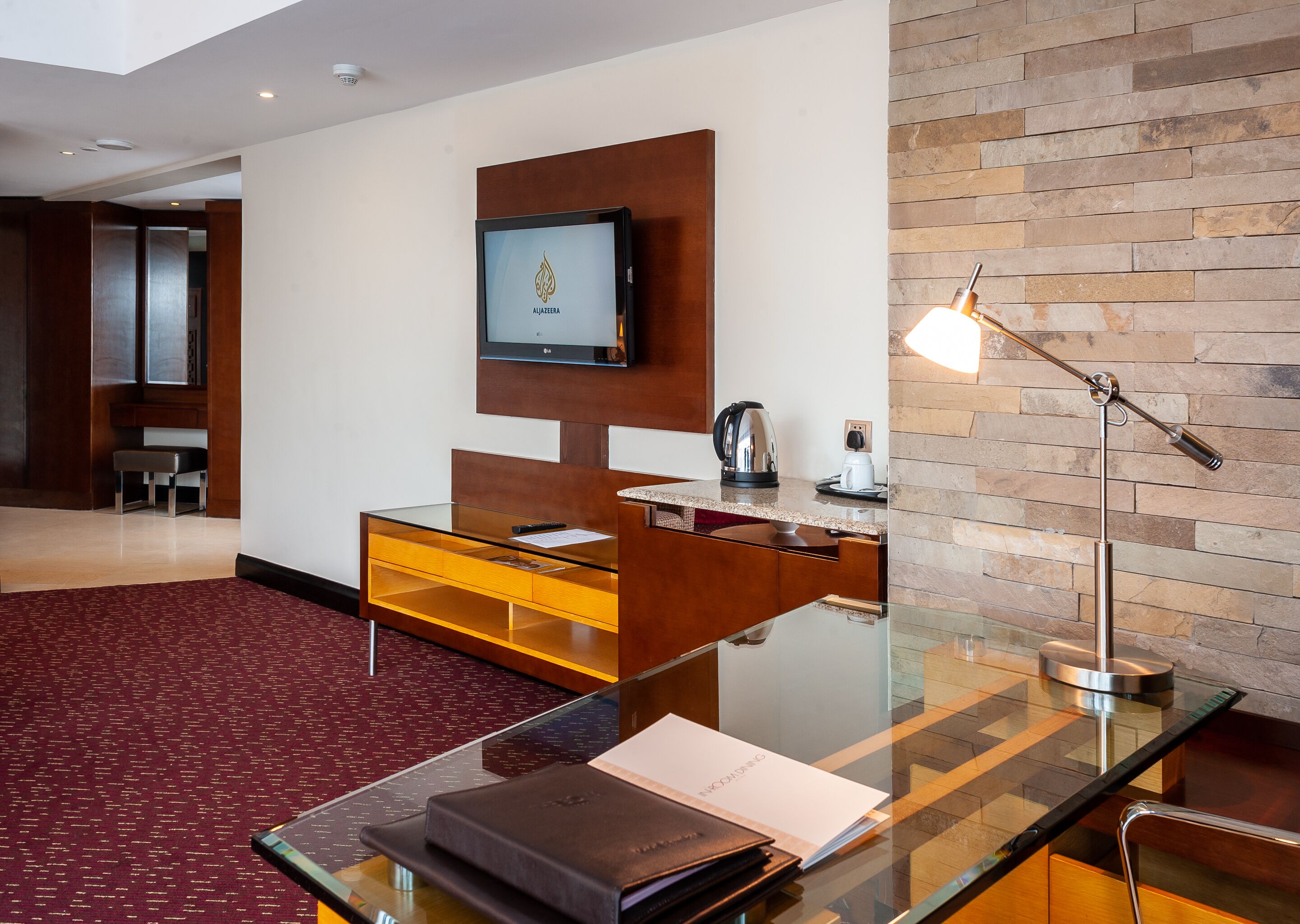 A modern hotel room with a glass desk, a flat-screen TV mounted on a wooden panel, a coffee maker, and a lamp.