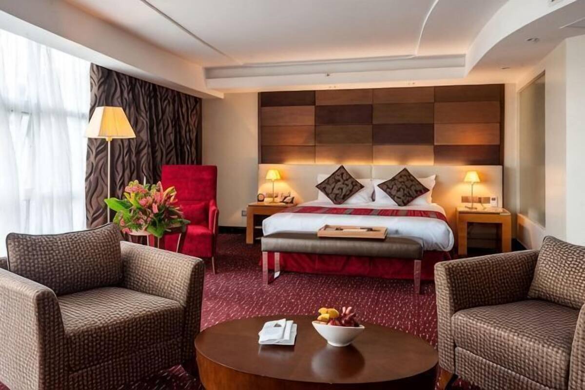 A hotel room with a bed, two bedside tables, a red chair, two armchairs, a coffee table, and a large window.