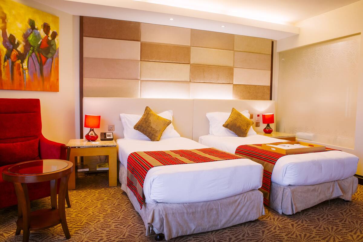 A hotel room with two beds, a red armchair, a small table, and a painting on the wall.