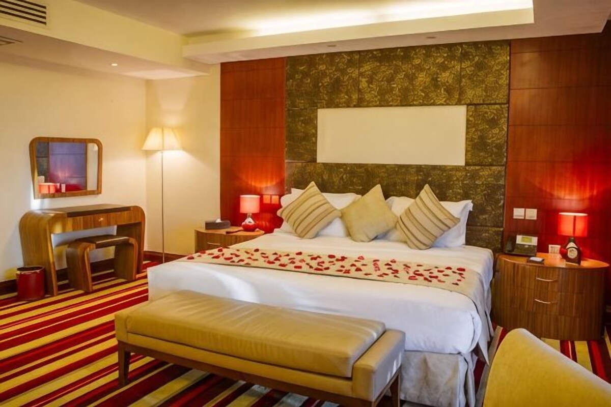 A hotel room with a large bed, a desk, a chair, a bench, and a nightstand with a lamp.