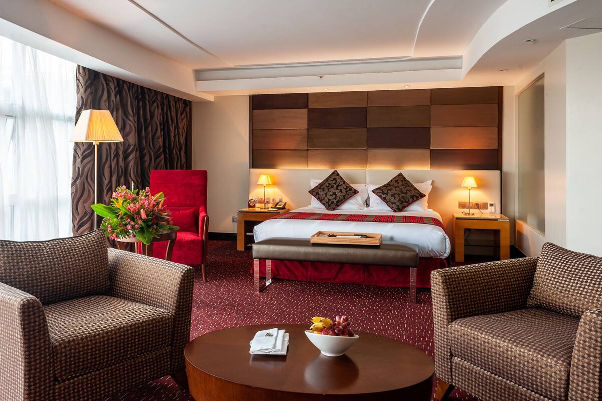 A hotel room with a large bed, two bedside tables with lamps, a red armchair, two armchairs, a round coffee table with a bowl of fruit, and a window with sheer curtains.