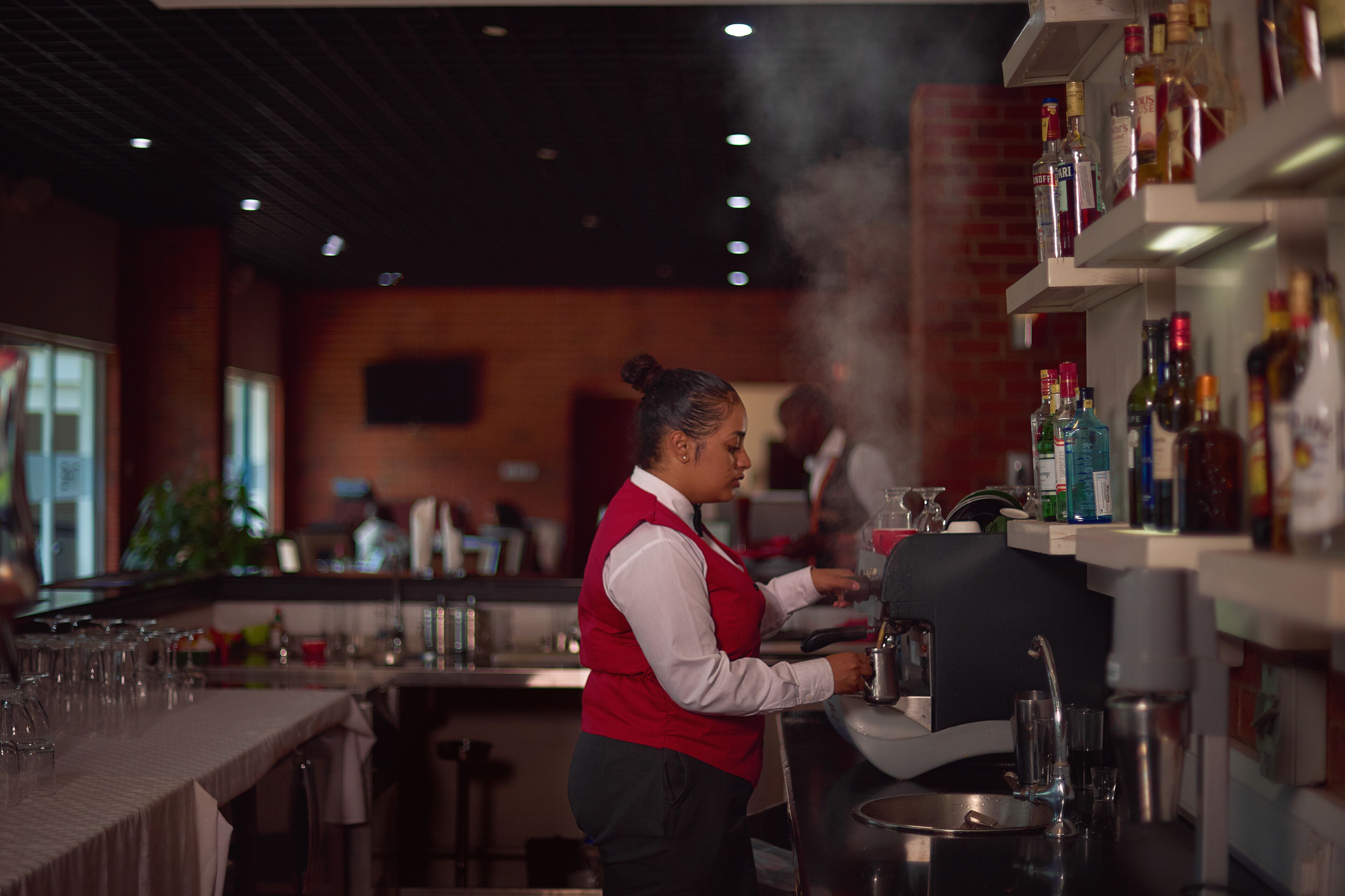Una zona de bar con un bartender preparando bebidas, estantes con botellas y una barra con vasos.