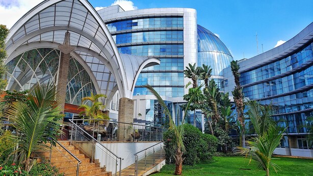 Un edificio moderno con fachada de vidrio curvo, un atrio central con techo de cristal y una escalera que conduce a un nivel superior.