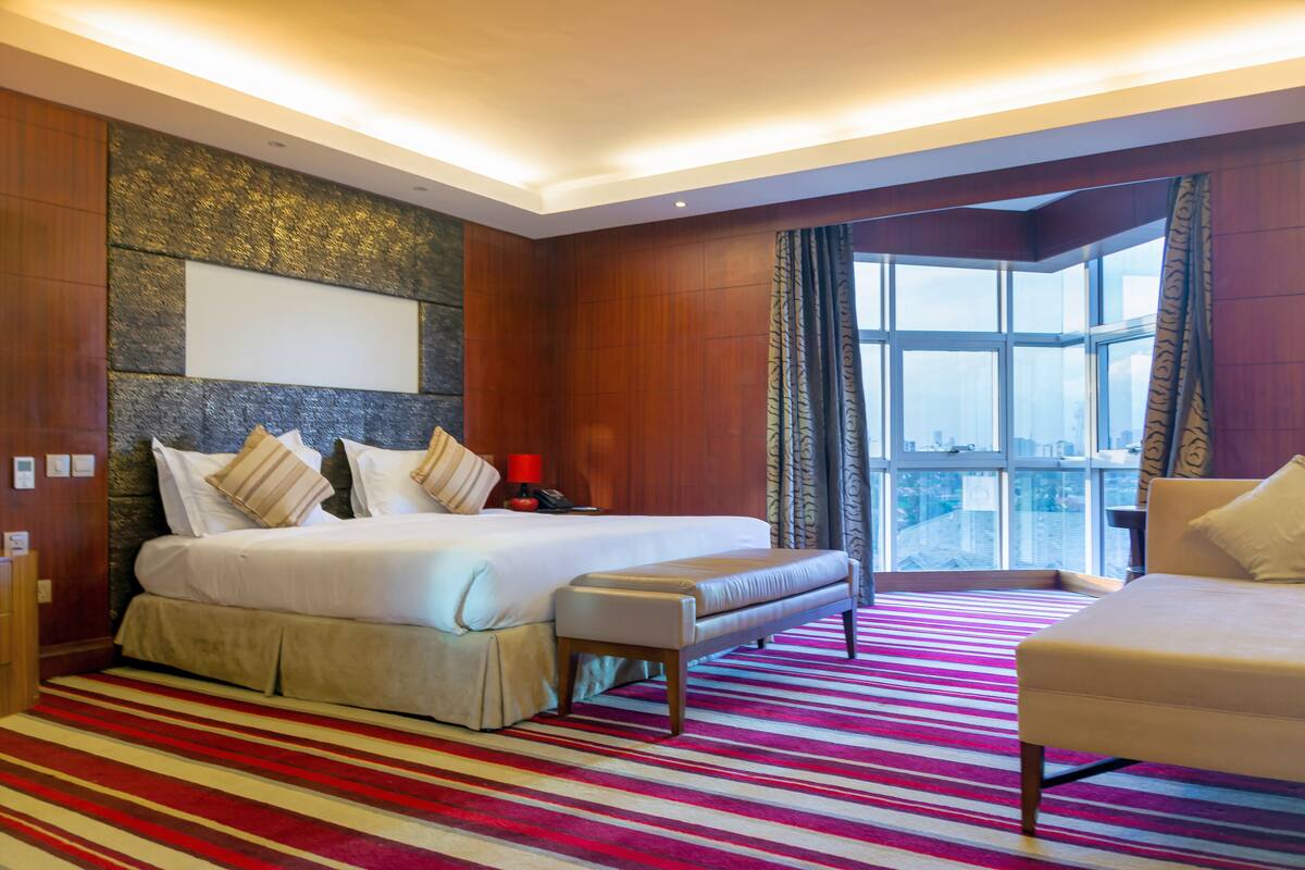 A hotel room with a large bed, a bench, a chair, a nightstand, and a view of the city through large windows.