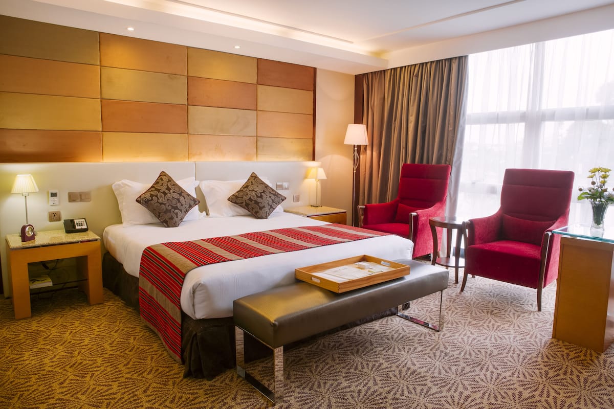 A hotel room with a large bed, red armchairs, a small table, and a bedside lamp.