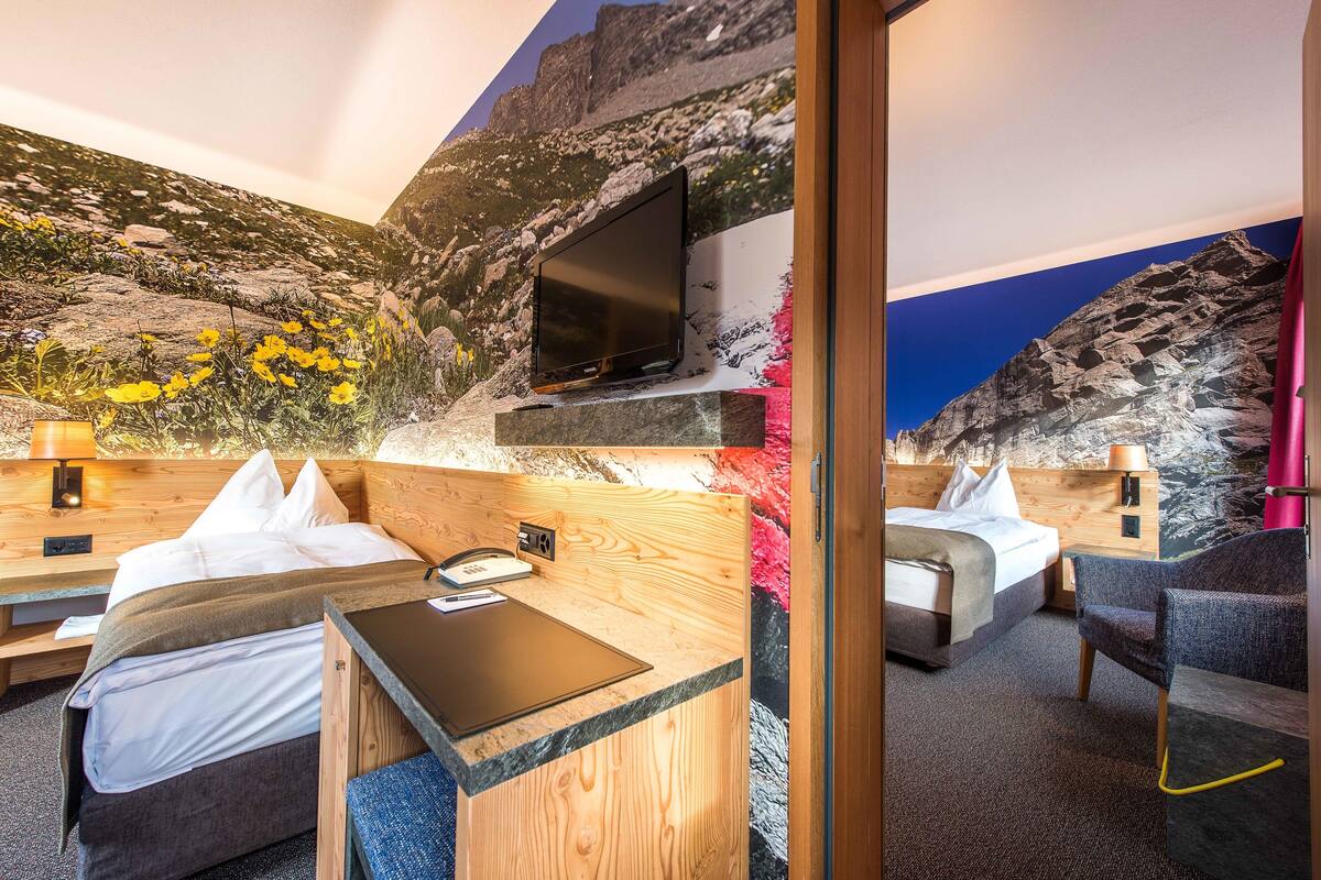 Habitación de hotel con cama, escritorio, silla, televisor y un mural de un paisaje montañoso.