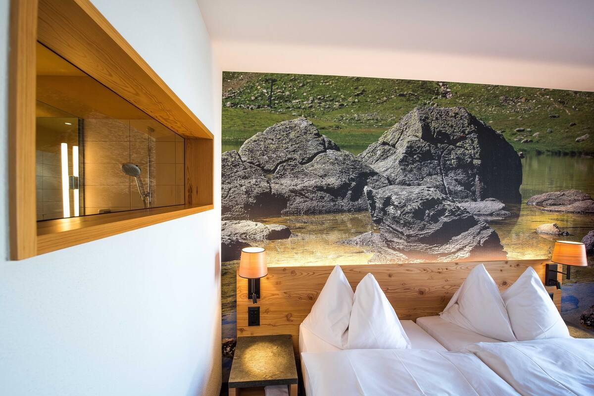 Un dormitorio con cama, mesita de noche, lámpara y un amplio mural en la pared que muestra un paisaje rocoso con un lago.