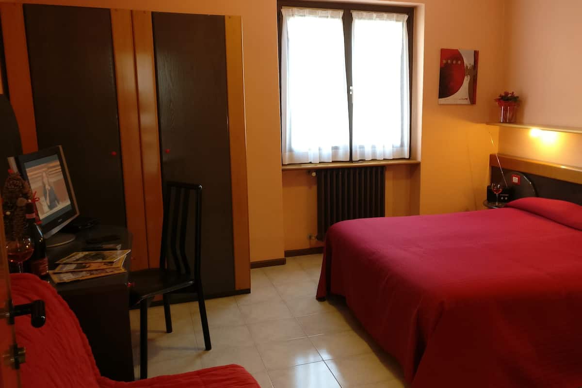 A hotel room with a bed, a desk with a computer, a chair, a window with curtains, a radiator, and a small table with a glass.
