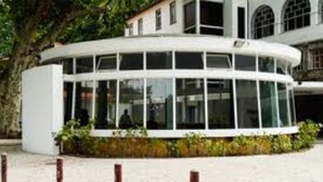 A modern building with a curved glass facade, surrounded by greenery and a sandy ground.