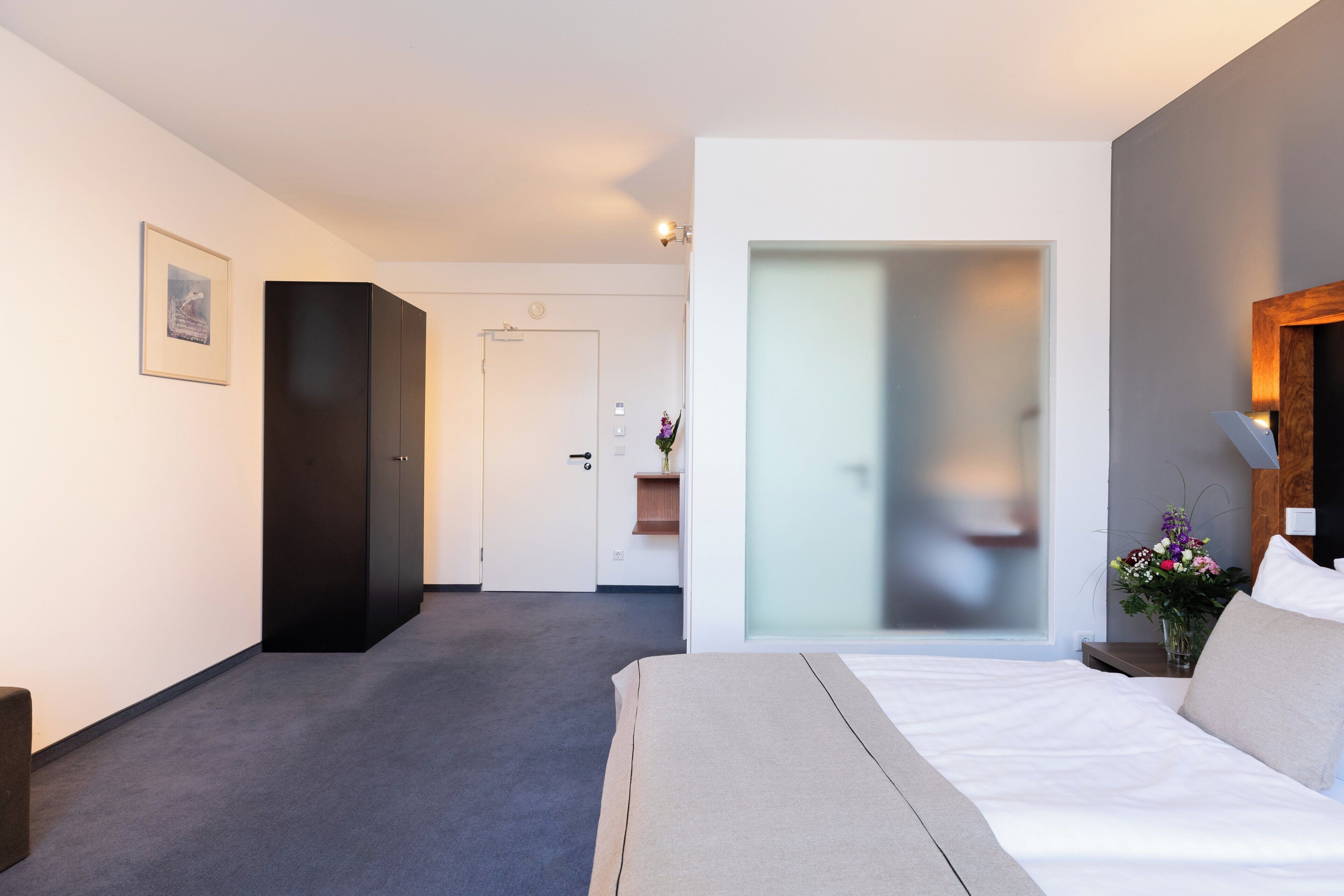 Una habitación de hotel moderna con una cama grande, un armario oscuro, una puerta que da a otra habitación y una mesita con un florero.