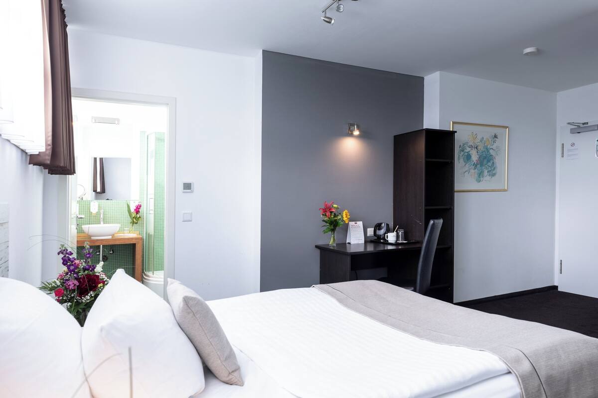 A hotel room with a bed, a desk, a chair, a small table with a vase of flowers, and a picture on the wall.