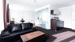 A modern living room with a black leather sofa, a wooden coffee table, and a glass partition leading to a kitchen area. - Hotel Aviva (Leipzig)