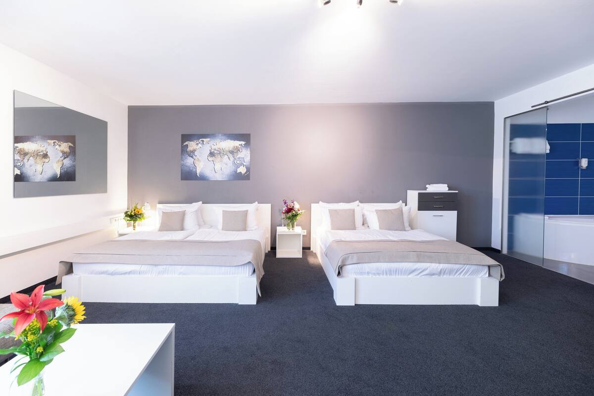 A modern hotel room with two beds, a small bedside table with a vase of flowers, a large world map on the wall, and a bathroom with blue tiles.