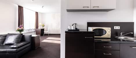 A modern hotel room with a black leather sofa, a compact kitchenette, and a large window with curtains.