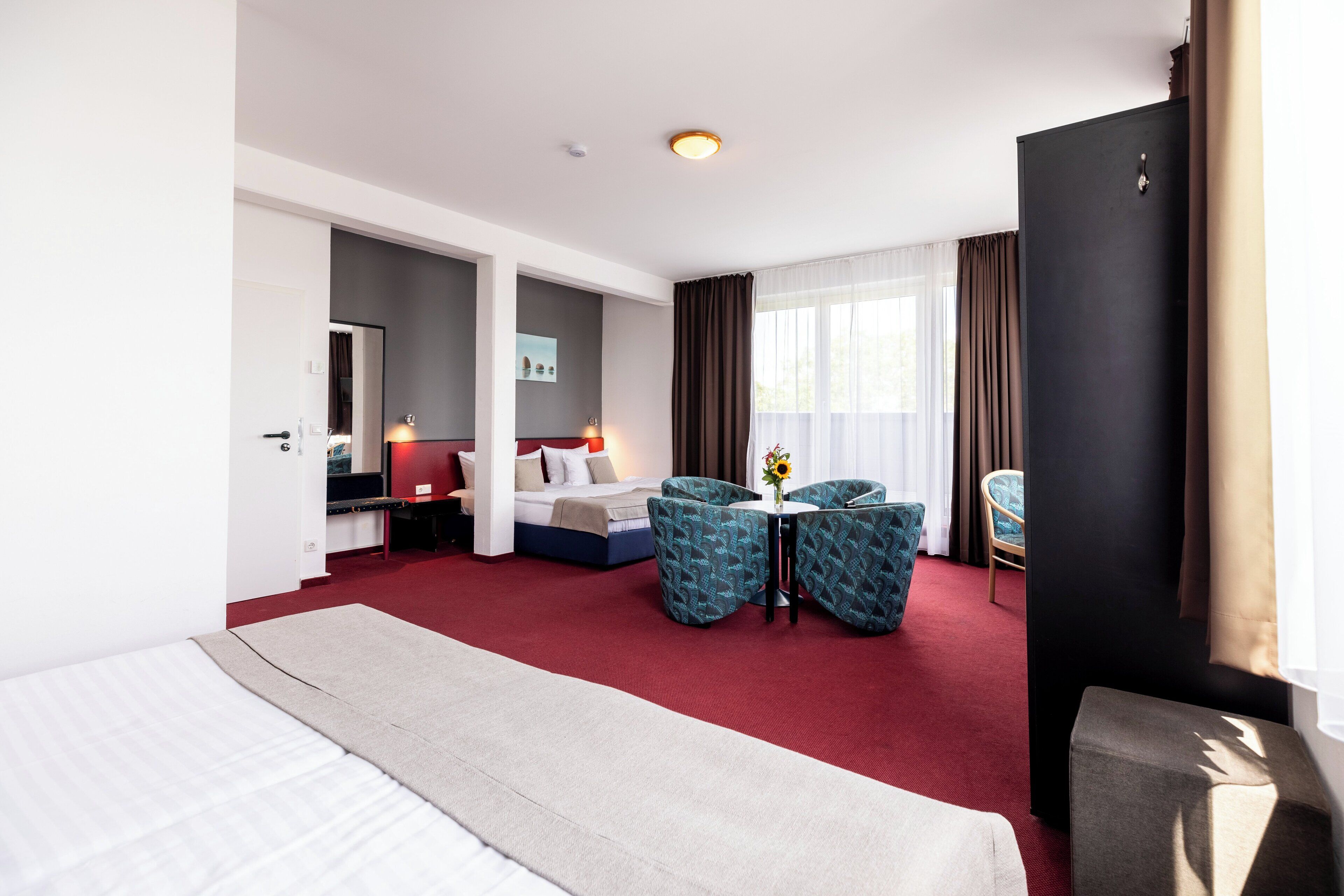 Habitación de hotel con una cama grande, un escritorio con una silla, dos sillones, una mesita con un florero y un ventanal con cortinas.