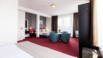 Habitación de hotel con una cama grande, un escritorio con una silla, dos sillones, una mesita con un florero y un ventanal con cortinas.