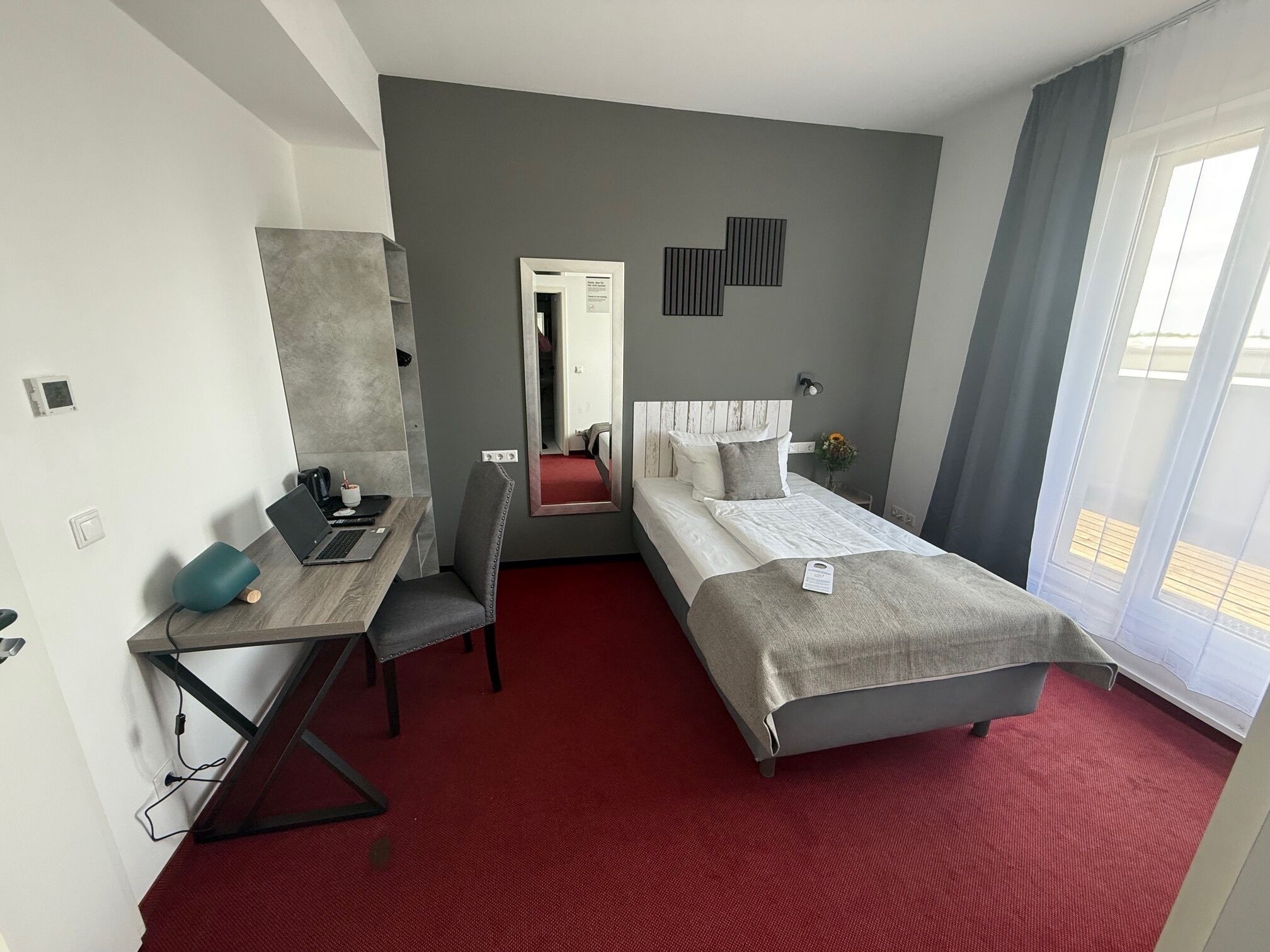 Ett hotellrum med en säng, ett skrivbord med en bärbar dator, en stol och en spegel.