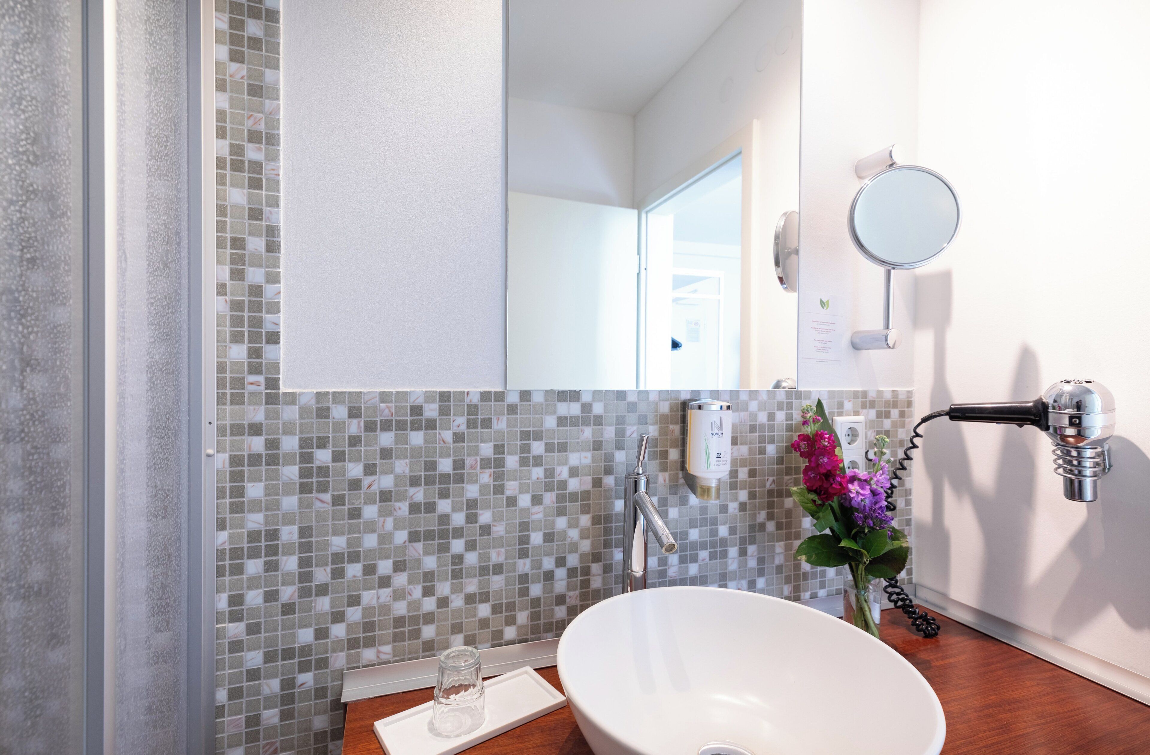 Uma casa de banho com uma pia branca, um espelho, um porta-toalhas, um frasco de vidro e um vaso de flores.