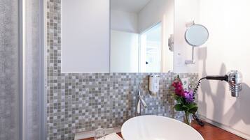 Uma casa de banho com uma pia branca, um espelho, um porta-toalhas, um frasco de vidro e um vaso de flores.