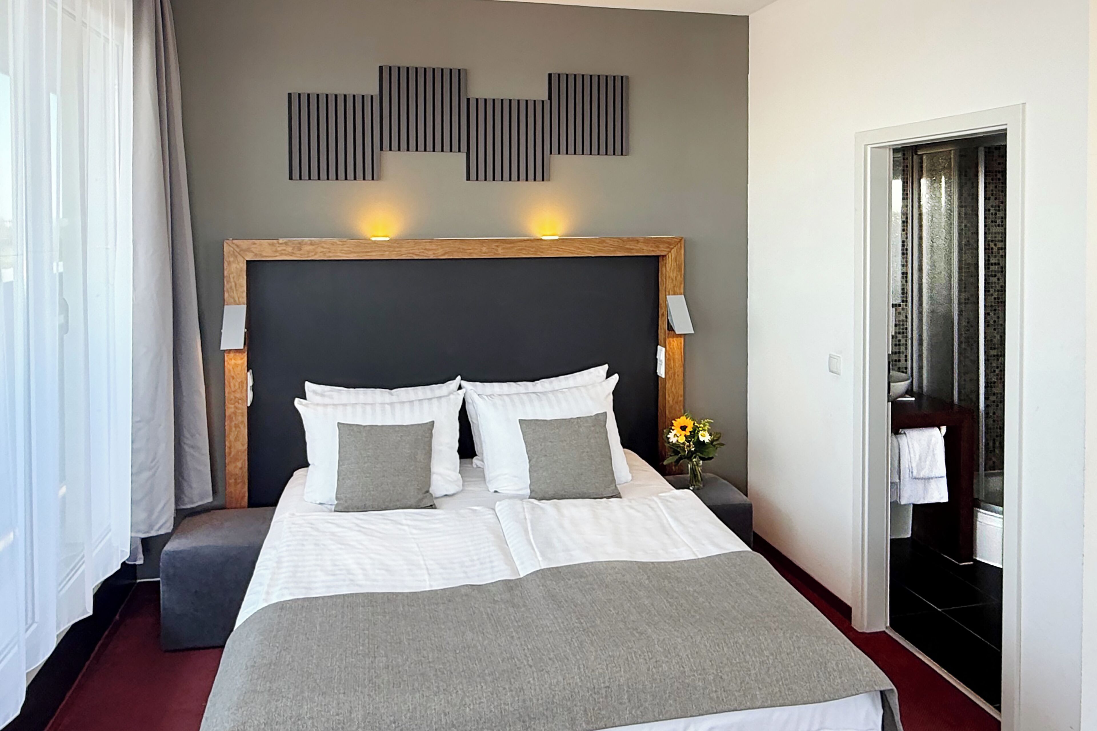 A hotel room with a large bed, a wooden headboard, a bedside table with a vase of flowers, and a view of the bathroom.