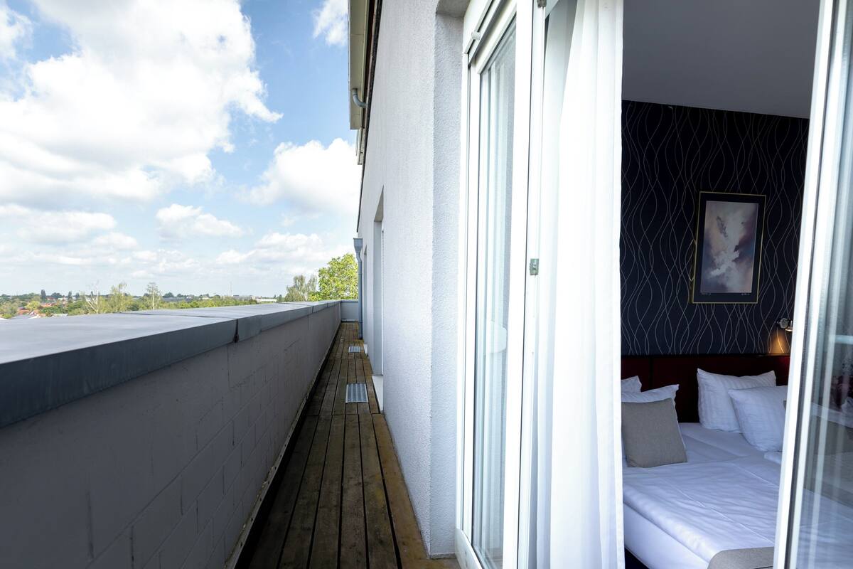 A hotel room with a balcony, a bed with white linens, a dark patterned wall, and a framed picture on the wall.