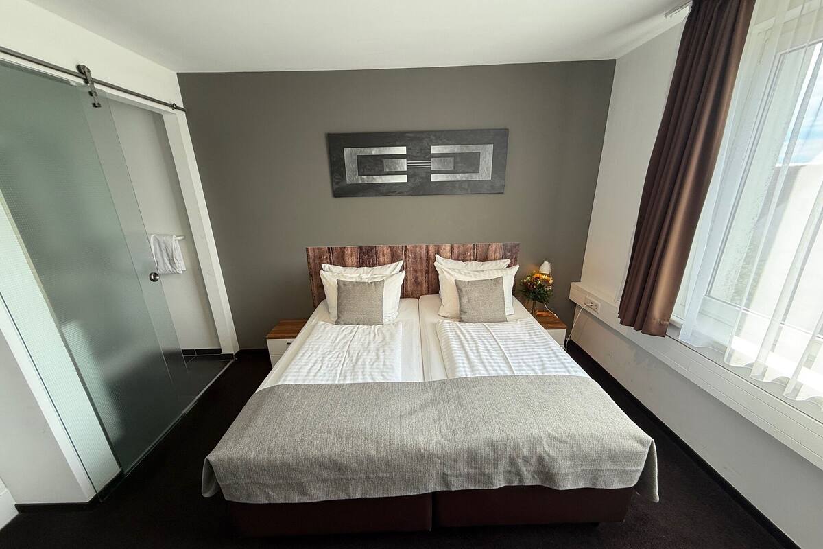 A hotel room with a large bed, a wooden headboard, a wall art piece, a window with curtains, and a door with a towel hanging on it.