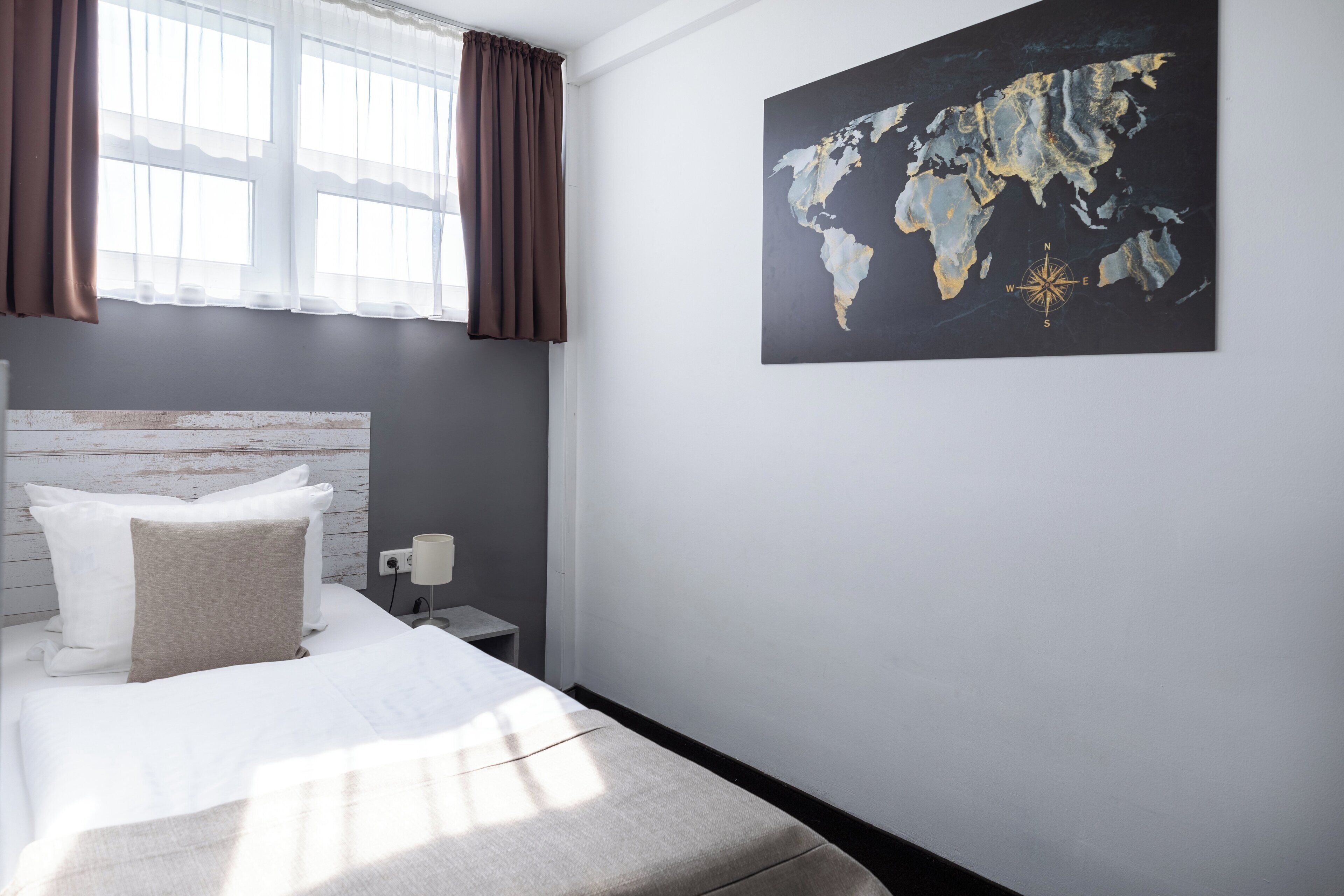 A neatly made bed with white linens and a beige throw, a small bedside table with a lamp, and a world map on the wall.