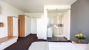 A modern hotel room with a large bed, a wooden wardrobe, a shower area, and a small bedside table with a vase of flowers. - Hotel Aviva (Leipzig)
