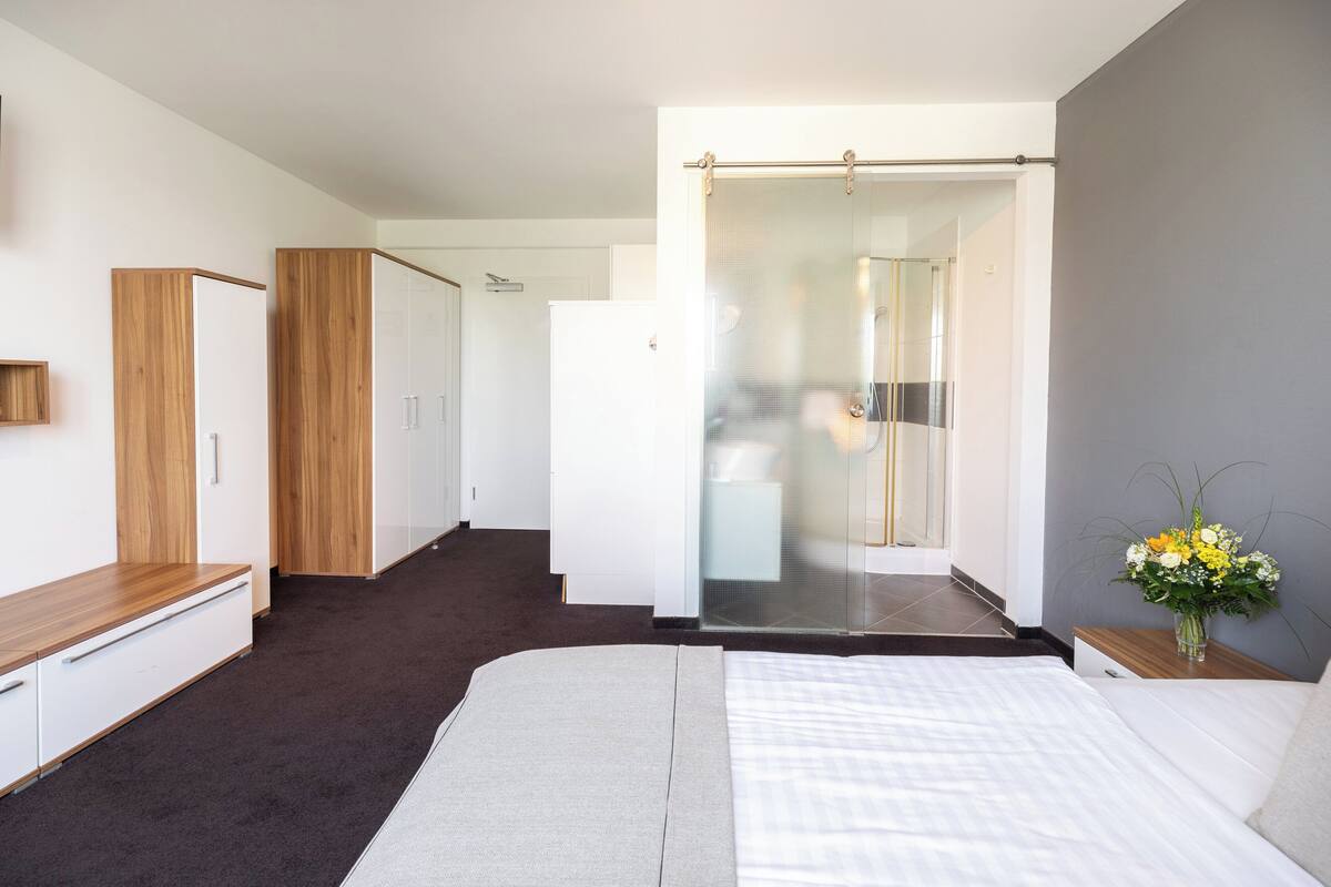 A modern hotel room with a large bed, a wooden wardrobe, a shower area, and a small bedside table with a vase of flowers.