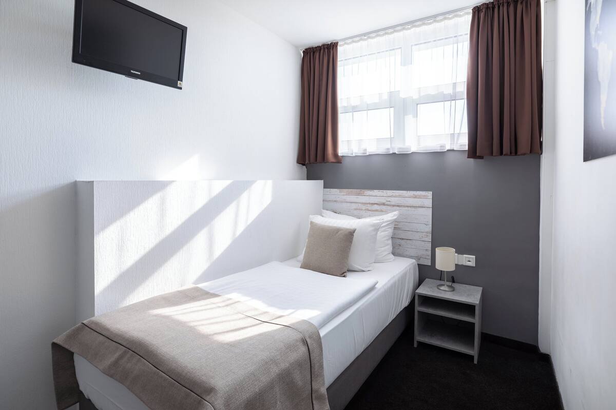 A small, neatly arranged bedroom with a single bed, a bedside table, a TV mounted on the wall, and a window with curtains.