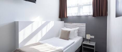Un dormitorio pequeño y ordenado con una cama individual, una mesita de noche, un televisor montado en la pared y una ventana con cortinas.