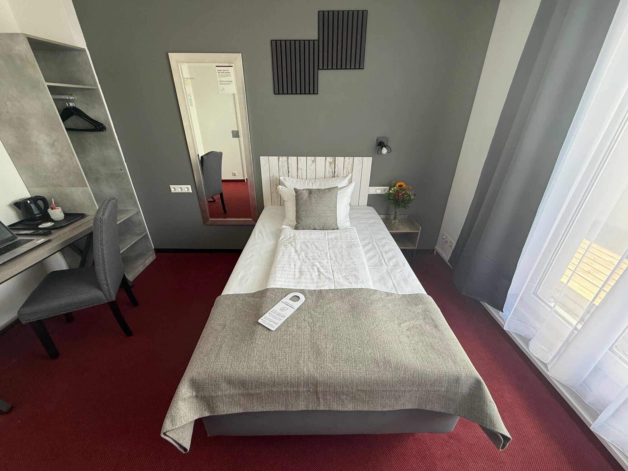 Quarto de hotel com cama, escrivaninha, cadeira, guarda-roupa, espelho e janela com cortinas.