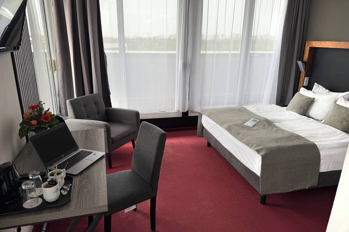 A hotel room with a bed, a desk with a laptop, a chair, and a dining area with a tray of food and drinks.