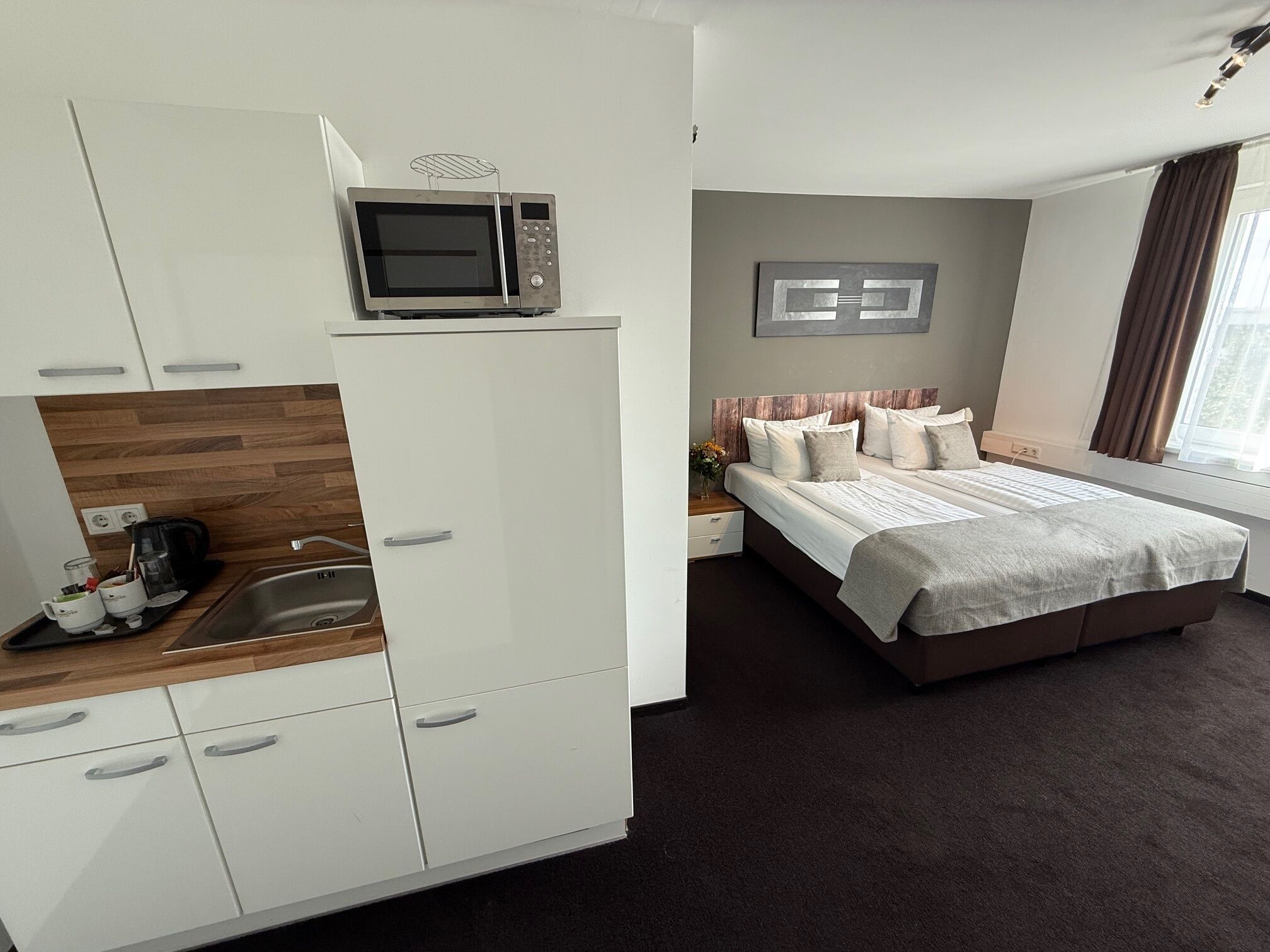 A modern hotel room with a kitchenette, a large bed with multiple pillows, and a view of the outdoors.