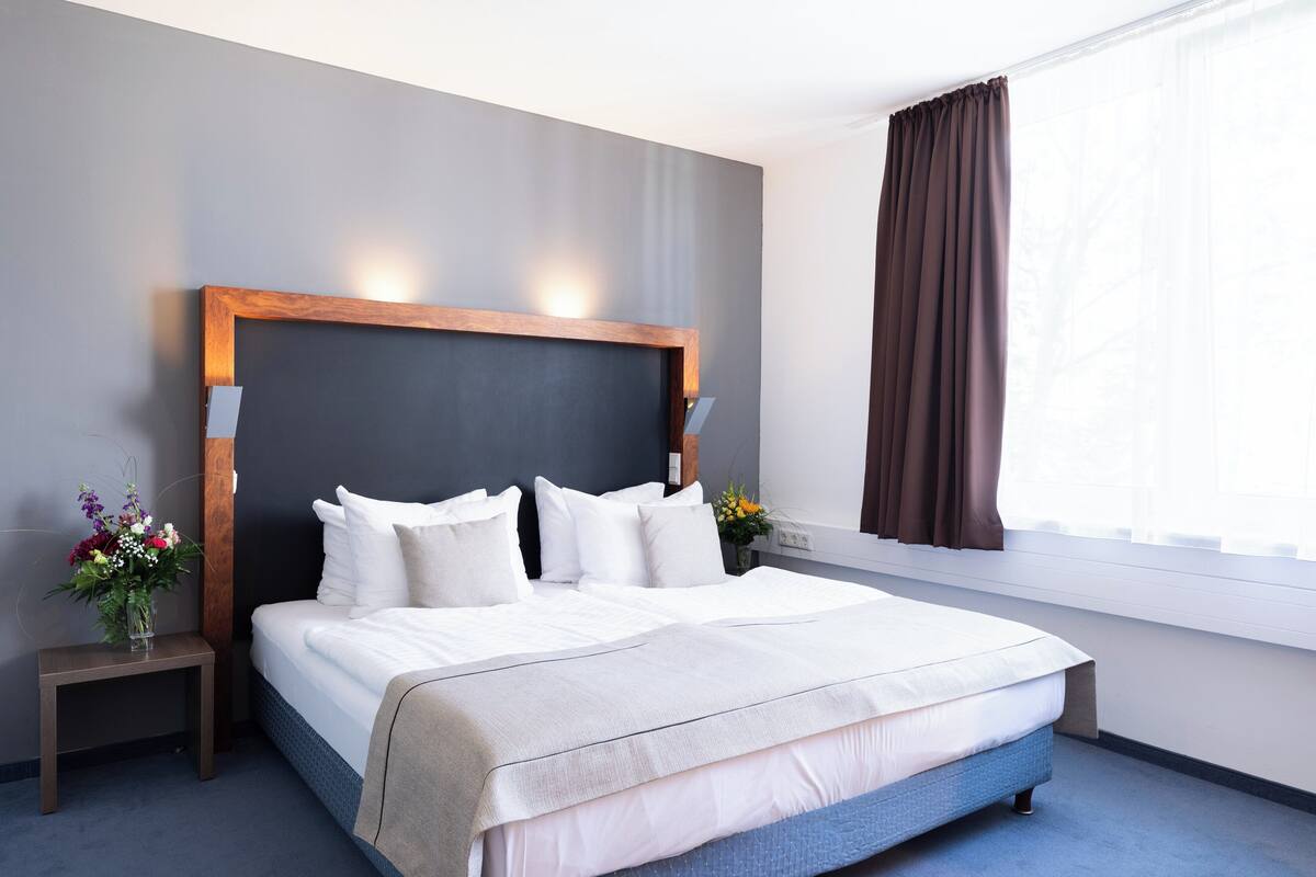 A hotel room with a large bed, a nightstand with a vase of flowers, and a window with curtains.