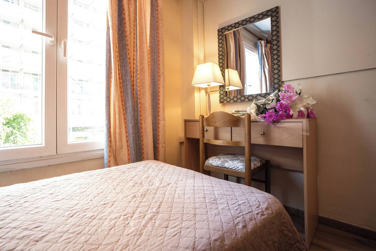 Una habitación de hotel con una cama, un tocador de madera con un espejo, una lámpara y un ramo de flores.