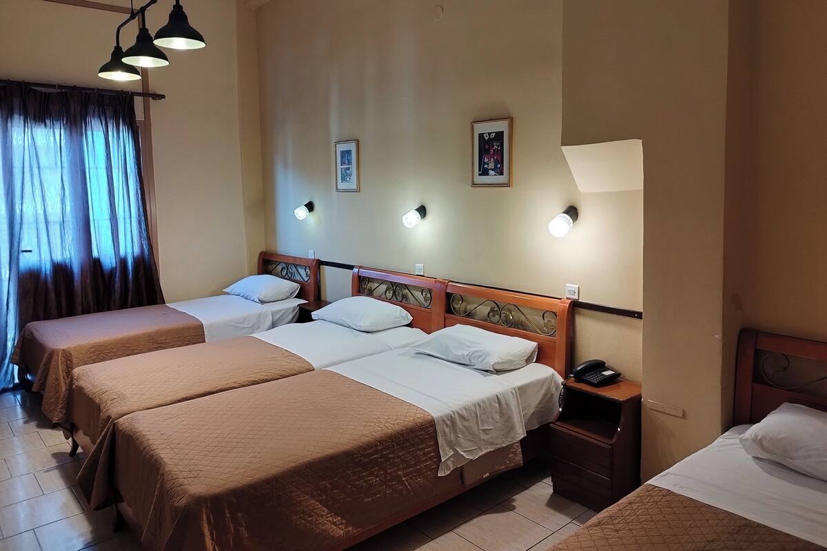 Habitación de hotel con tres camas, cada una con cabecero de madera y ropa de cama blanca. Hay luces fijadas en la pared y una mesita de noche pequeña con un teléfono.