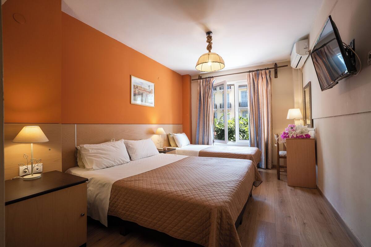 Habitación de hotel con dos camas, una mesita de noche con lámpara, un televisor montado en la pared y una ventana con cortinas.