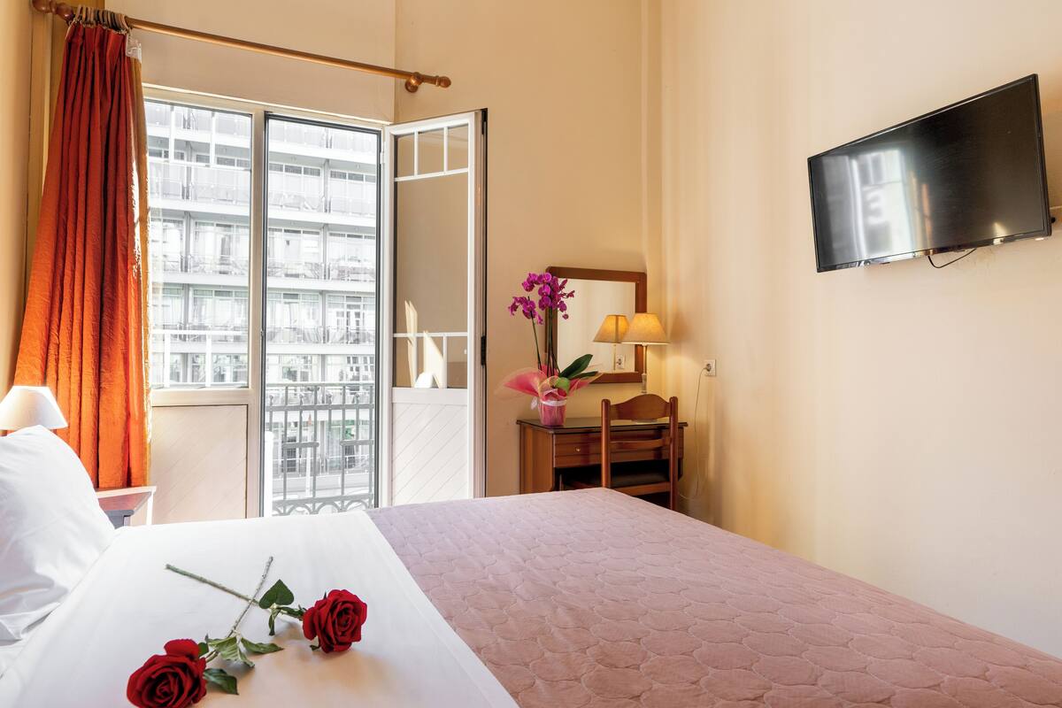 Habitación de hotel con cama, televisor montado en la pared, escritorio con lámpara y vistas a la ciudad a través de la ventana.