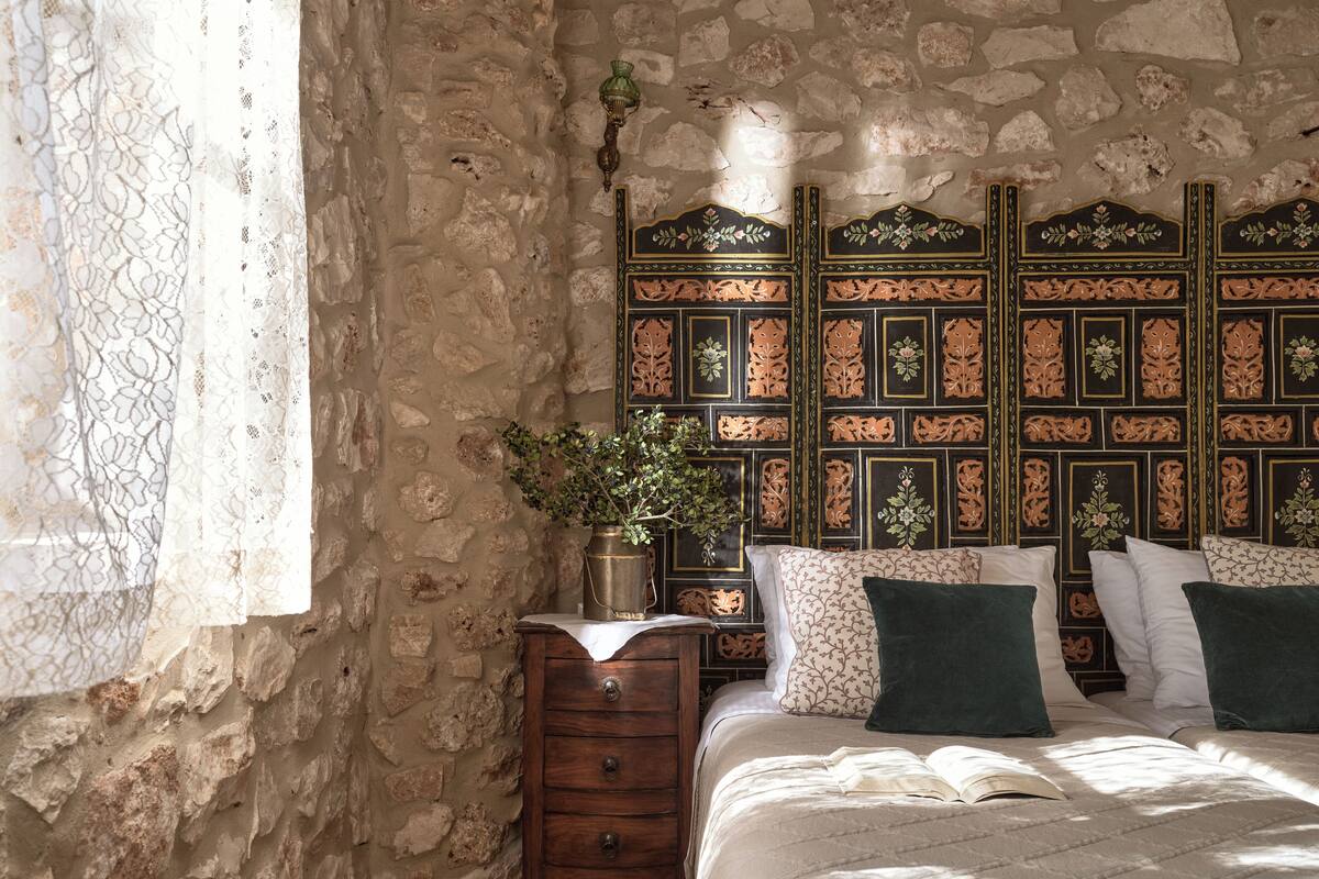 A bedroom with a stone wall, a wooden nightstand, a bed with patterned pillows, and a window with lace curtains.