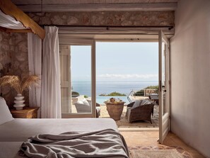A bedroom with a bed, a nightstand, a vase with dried plants, and a view of the sea through an open door. - Orfos Villas (Zakynthos)
