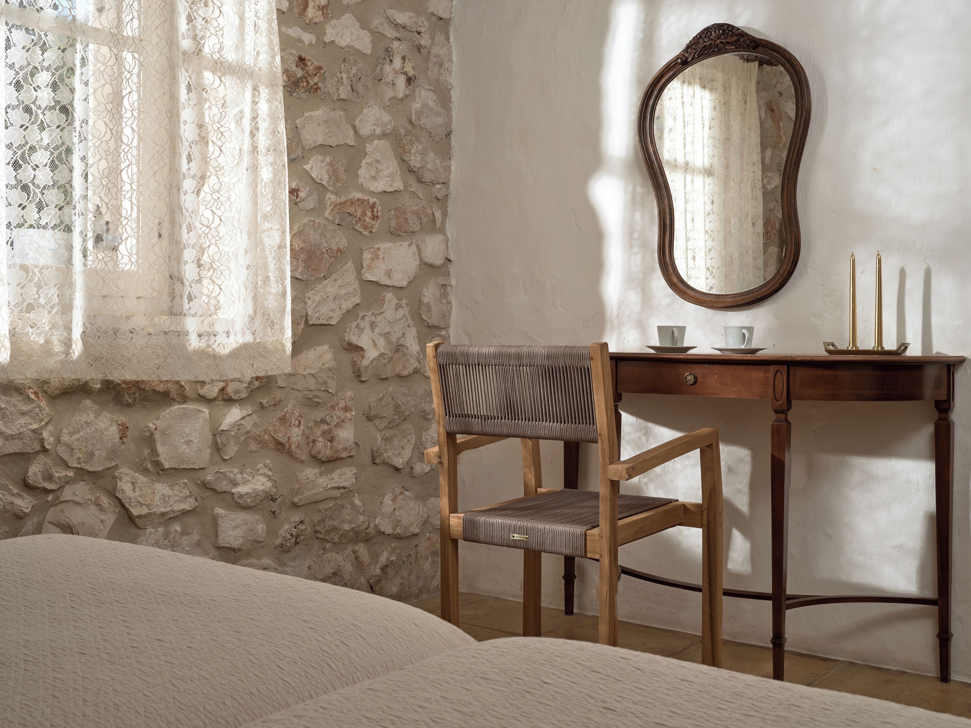 Una habitación con pared de piedra, un escritorio de madera con espejo, una silla y una cama.