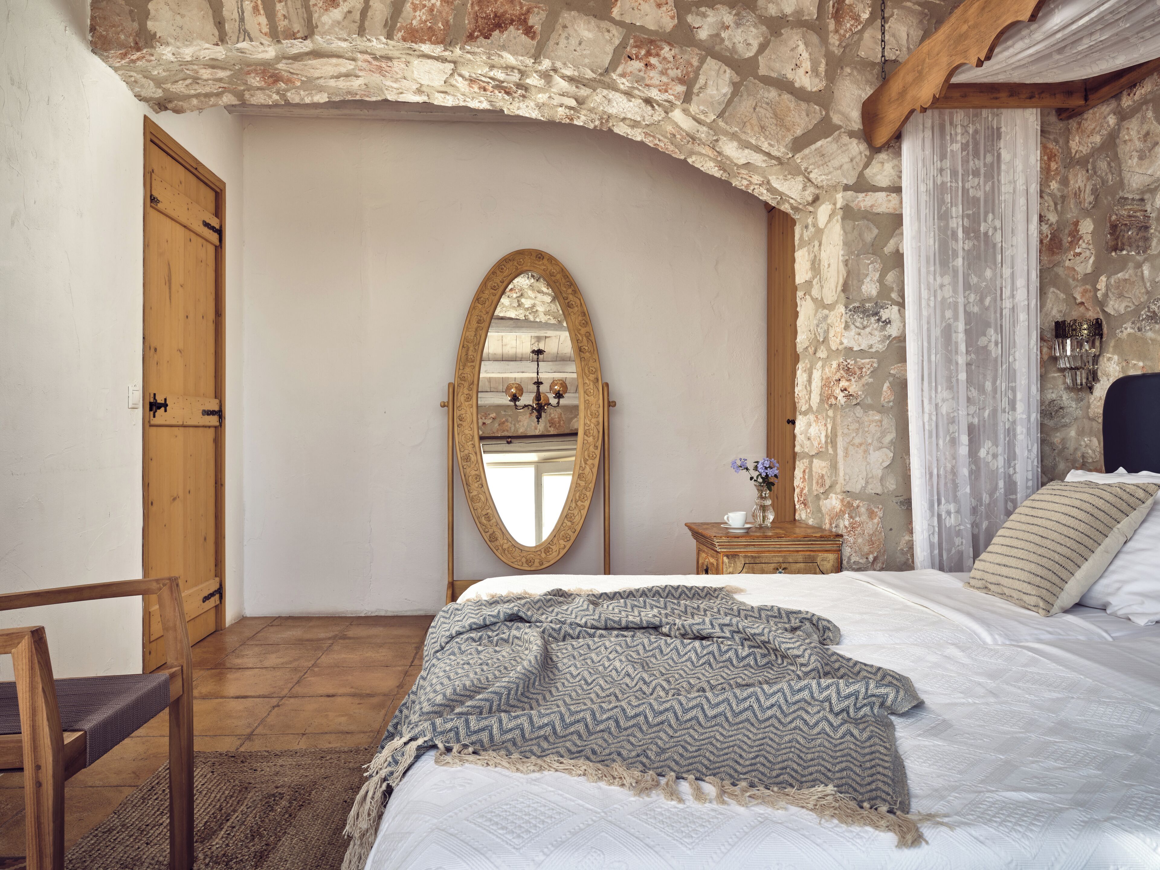 A bedroom with a stone wall, a wooden door, a bed with a patterned blanket, a bedside table with a vase, and a mirror.