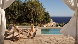 A wooden lounge chair with a checkered cushion, a stone pool, and a view of the sea.
