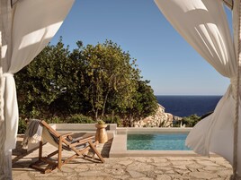 Una silla de salón de madera con cojín de cuadros, una piscina de piedra y vista al mar.